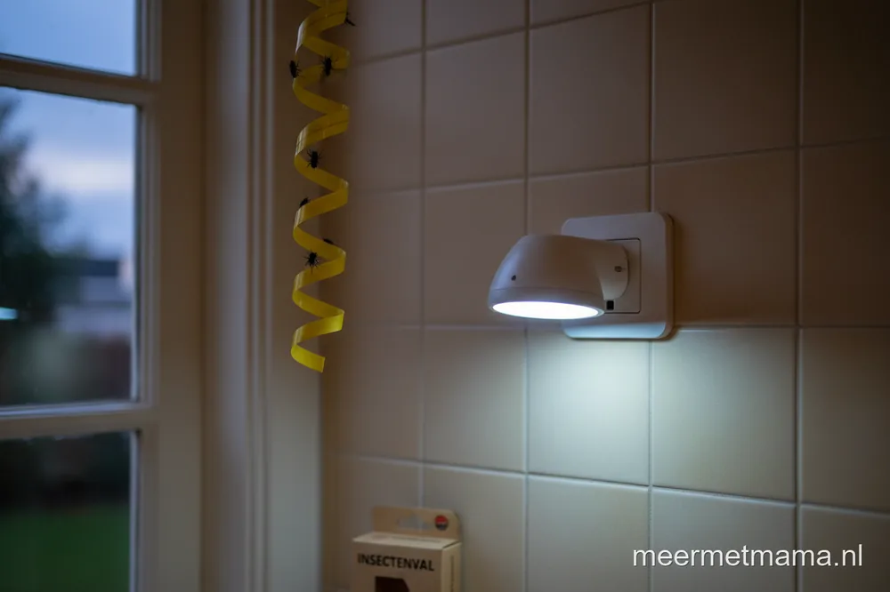 Een witte insectenlamp met blauw licht in het stopcontact, met op de achtergrond een gele plakrol met vliegen.